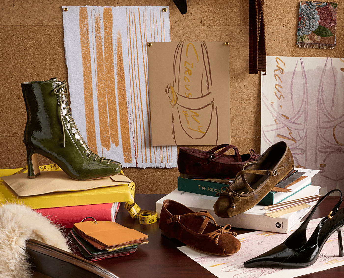 A design studio desk displaying Circus NY shoes: a green boot, black slingback pump, and suede Mary Jane flats.