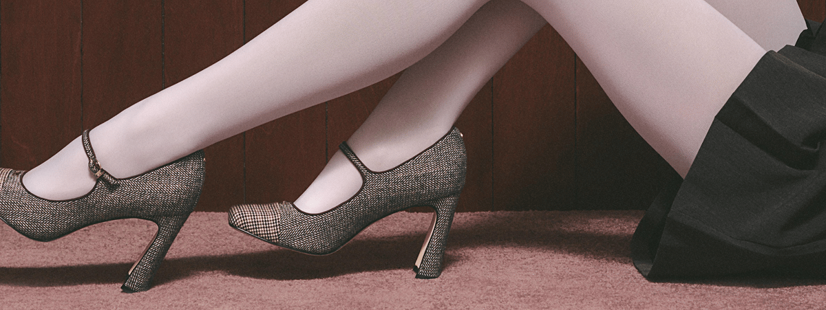 On-foot view of textured metallic Mary Jane pumps with a sculpted heel, styled with white tights and a black skirt.