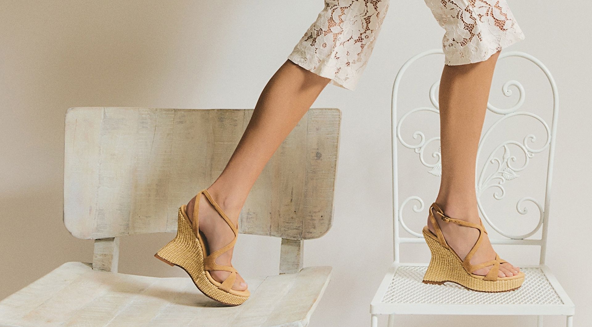 Model standing on white chairs wearing the Trina platform wedge sandal with a woven platform wedge, styled with white lace knee-length shorts.