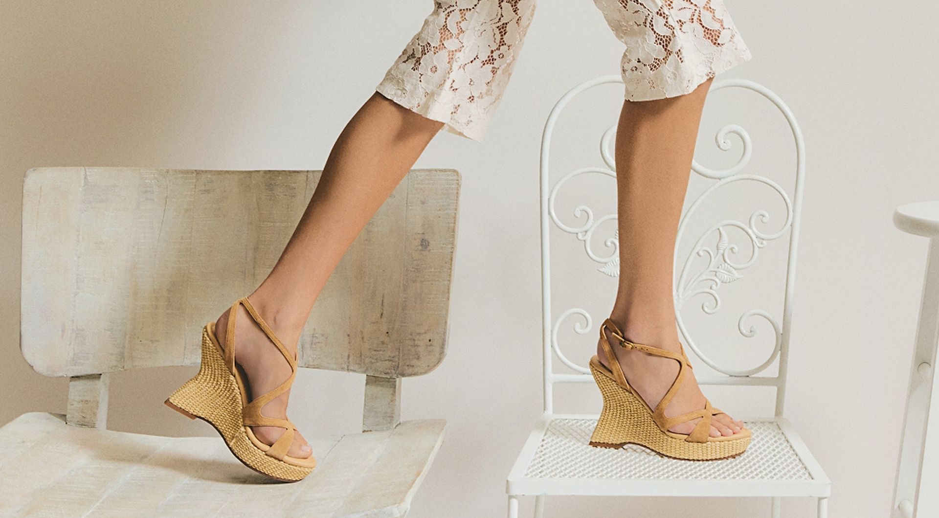 Model standing on white chairs wearing the Trina platform wedge sandal with a woven platform wedge, styled with white lace knee-length shorts.