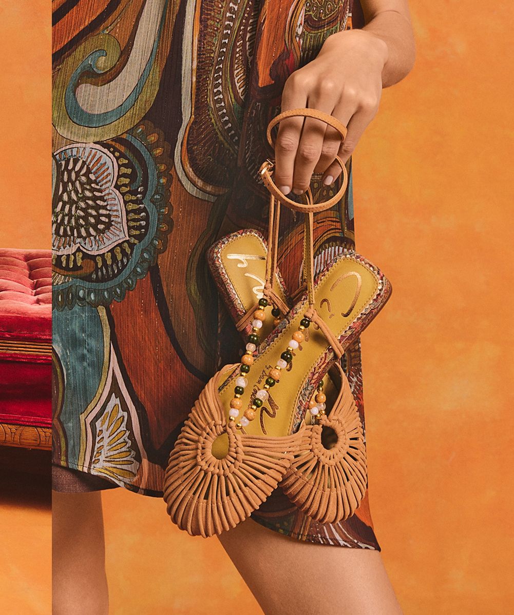 Model holding the Kassie ankle strap flat in amber sand suede with a woven fan-toe detail and beaded ankle strap.