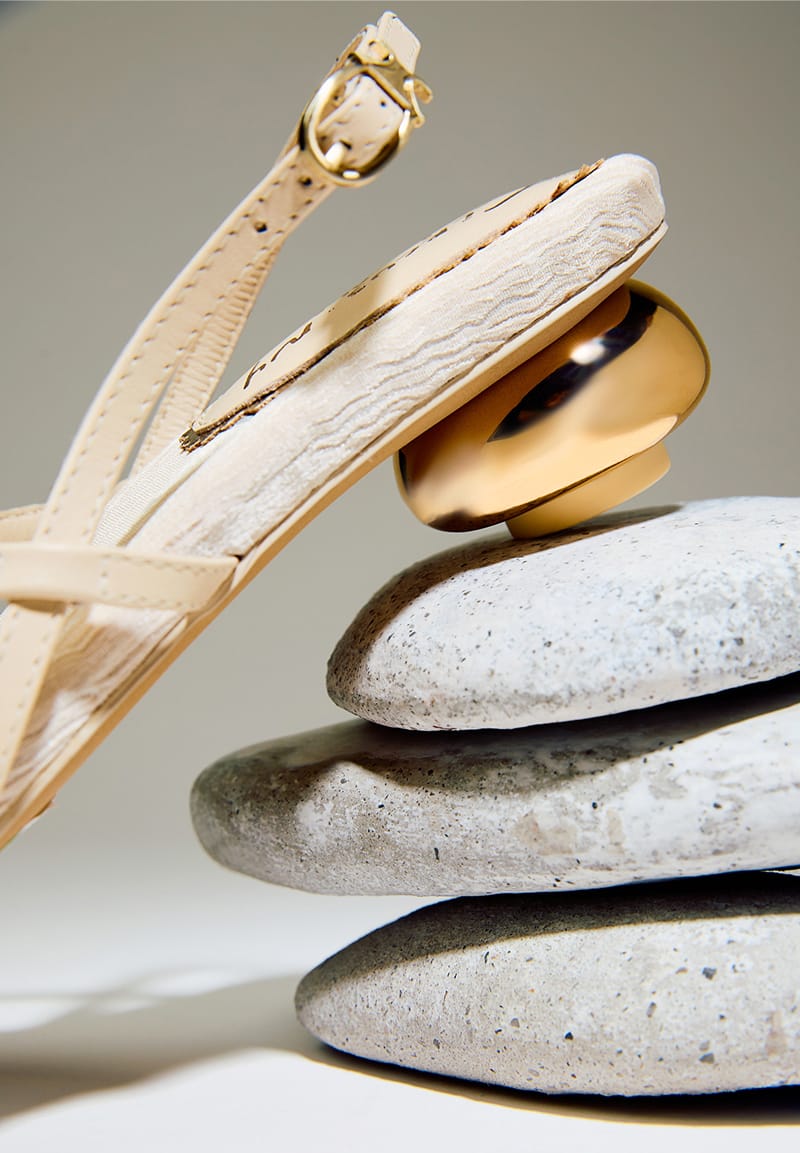 Circus NY Caia Heeled Sandal in Vanilla Bean Leather with a gold sculptural heel, resting on smooth grey river stones.